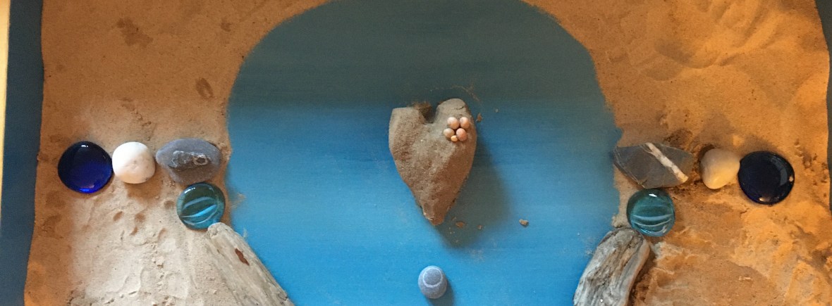 sand heart created in centre of tray blue background with shells, glass beads, pebbles and driftwood